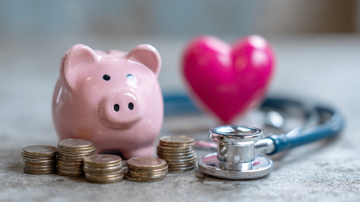 Pink piggy bank next to stacks of coins, a stethoscope, and a heart shape, symbolizing healthcare costs and insurance coverage
